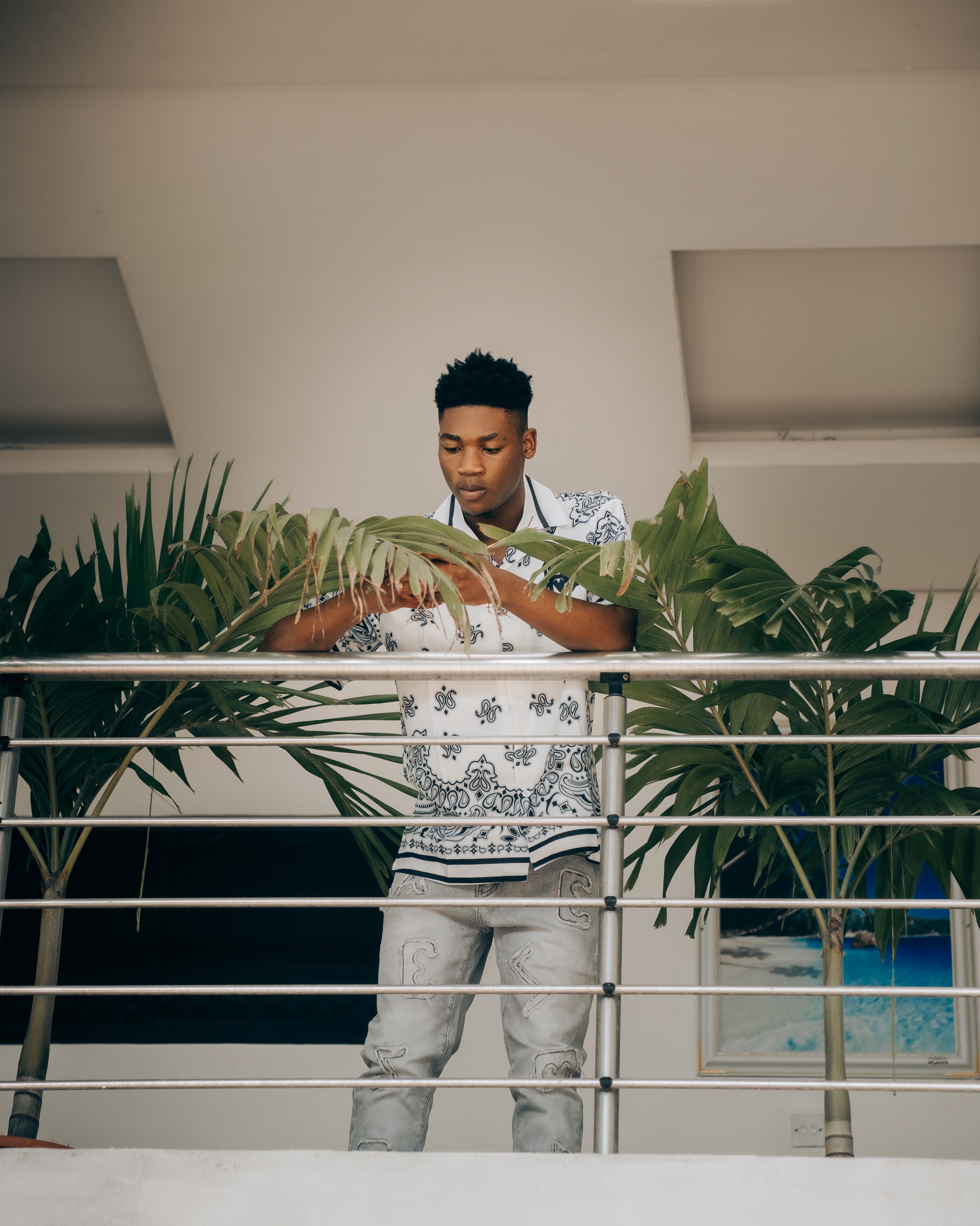 Young man in fashionable attire standing on a balcony surrounded by greenery indoors.