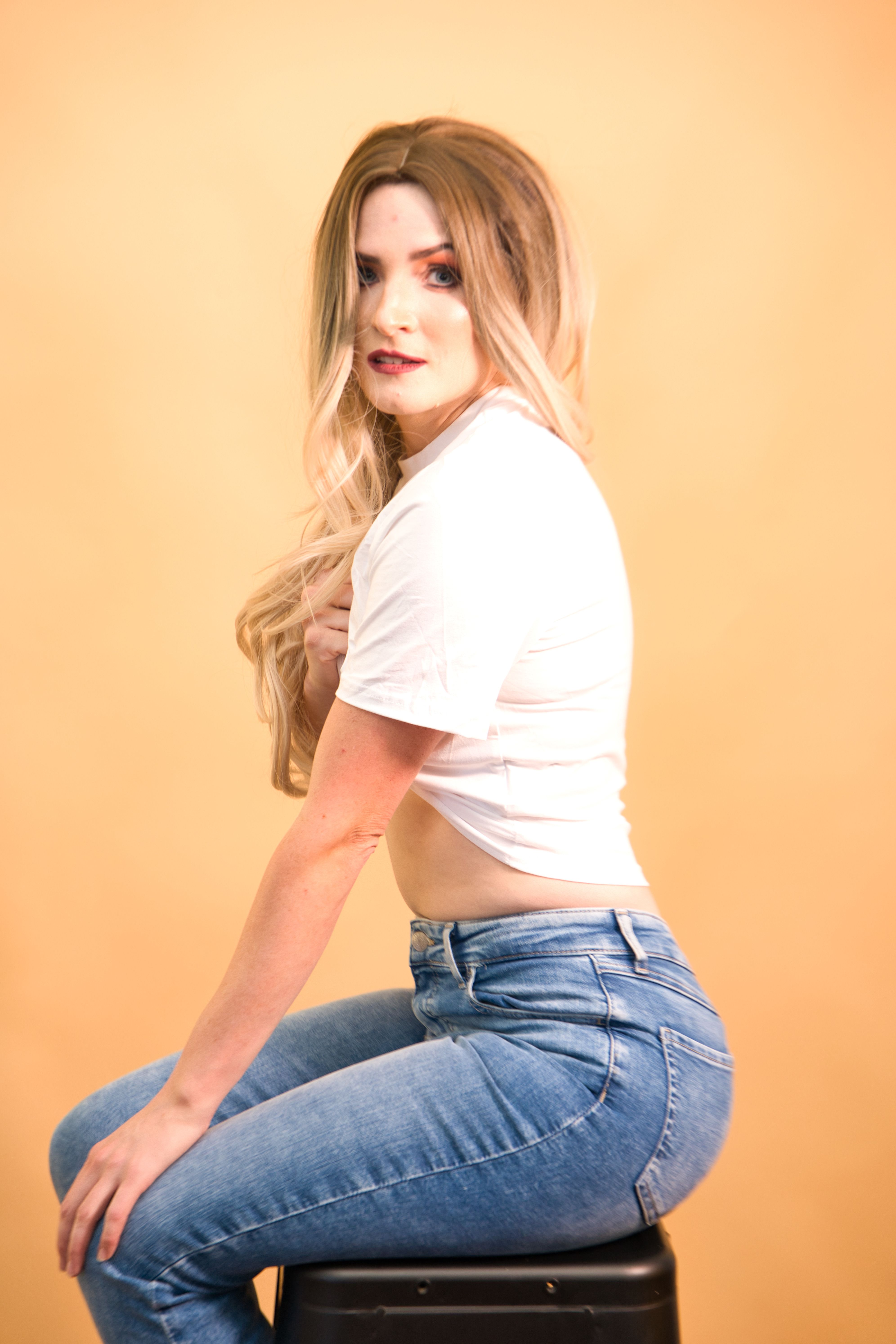 Stylish portrait of a young woman in jeans sitting on a stool against a beige background.