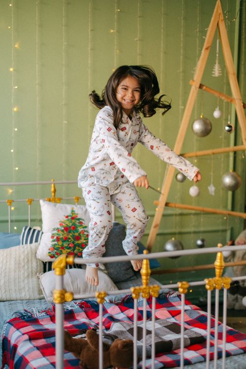 Free A young girl in pajamas excitedly jumps on a bed by a holiday-decorated room. Stock Photo