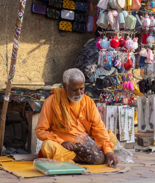 Free Elderly man in orange sitting at a street market in Vrindavan, India, selling colorful items. Stock Photo