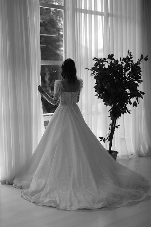 Black and White Photograph of a Bride Standing by a Window with Curtains