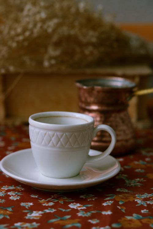 Free Elegant porcelain cup on saucer with a copper coffee pot in the background, perfect for a warm and cozy breakfast setup. Stock Photo