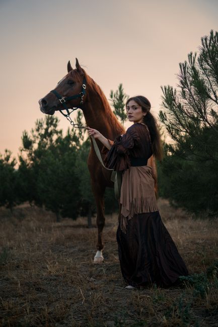 A woman stands by a horse amidst trees during sunset in Baku, Azerbaijan.