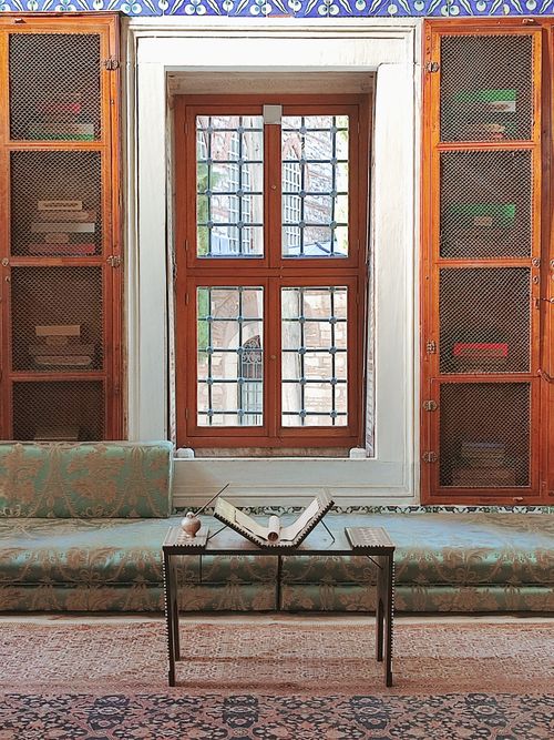 Free Beautifully preserved vintage room with an open Quran on a small table by a detailed window and wooden shelves. Stock Photo