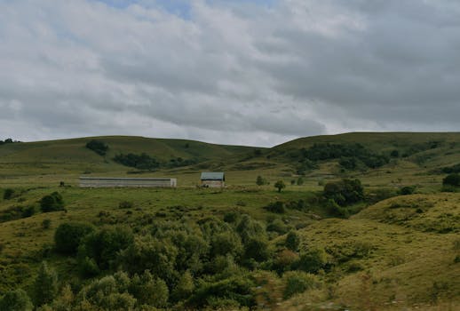 A tranquil countryside landscape featuring rolling hills and farm buildings under a cloudy sky.