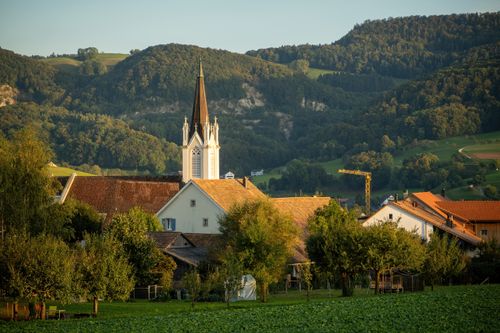 Free Quaint village church with spire surrounded by lush green hills and trees, bathed in daylight. Stock Photo