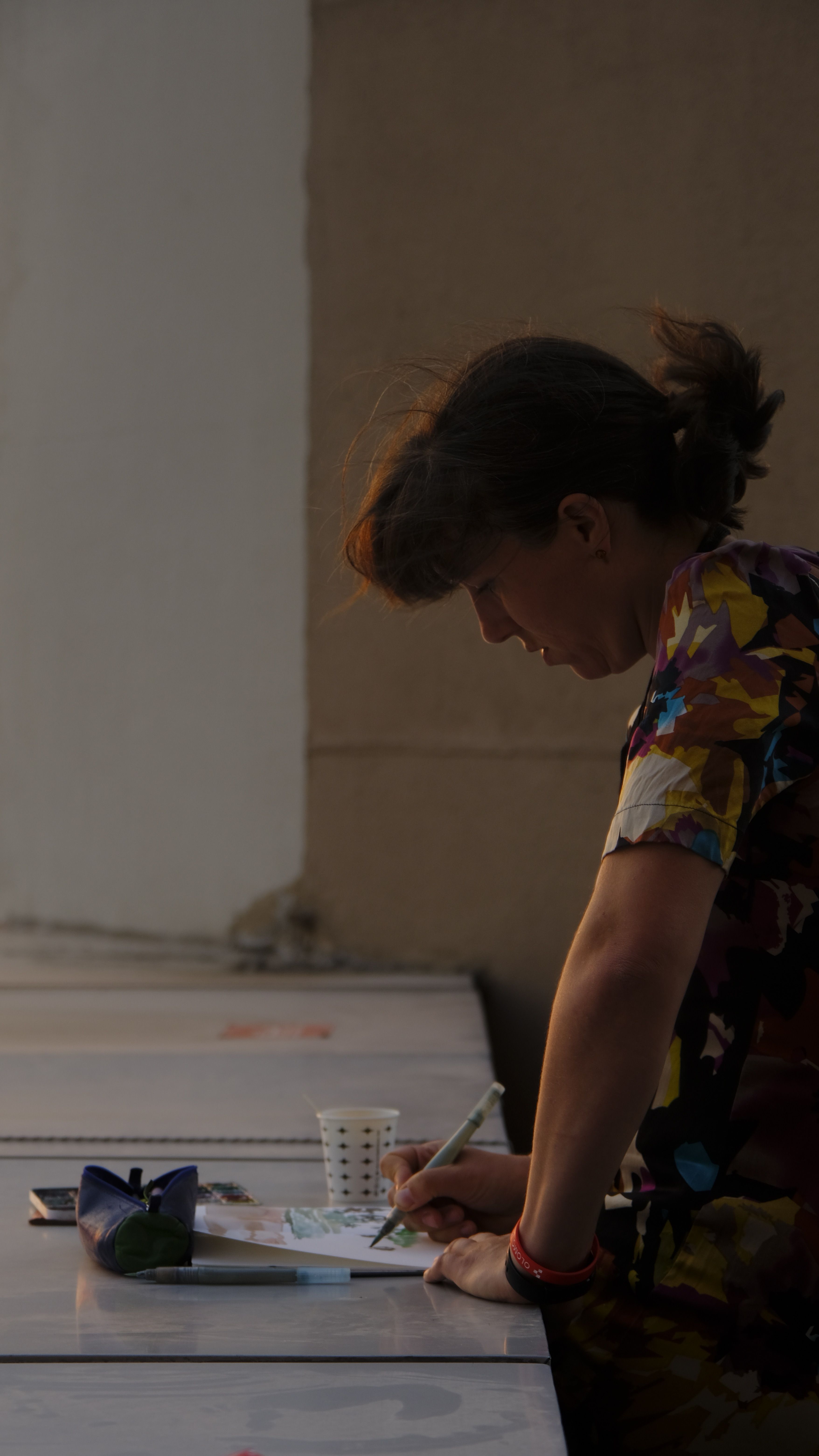 A woman sketches at an outdoor table in warm evening light.