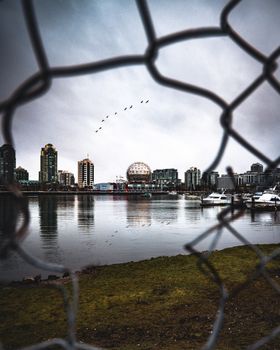 Dramatic view of Vancouver skyline through chain link fence with reflections on the water.