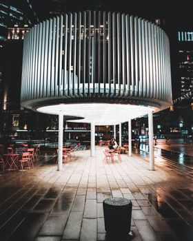 A sleek, modern urban pavilion illuminated at night with cityscape in background.