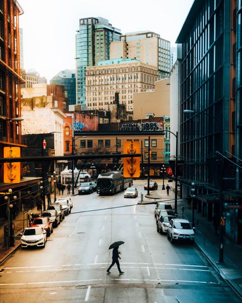 Free Rainy city crosswalk with buildings, cars, and a person walking under an umbrella in evening light. Stock Photo