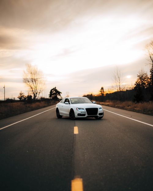 Free A stylish white car travels down a clear road at sunset, surrounded by trees and open sky. Stock Photo