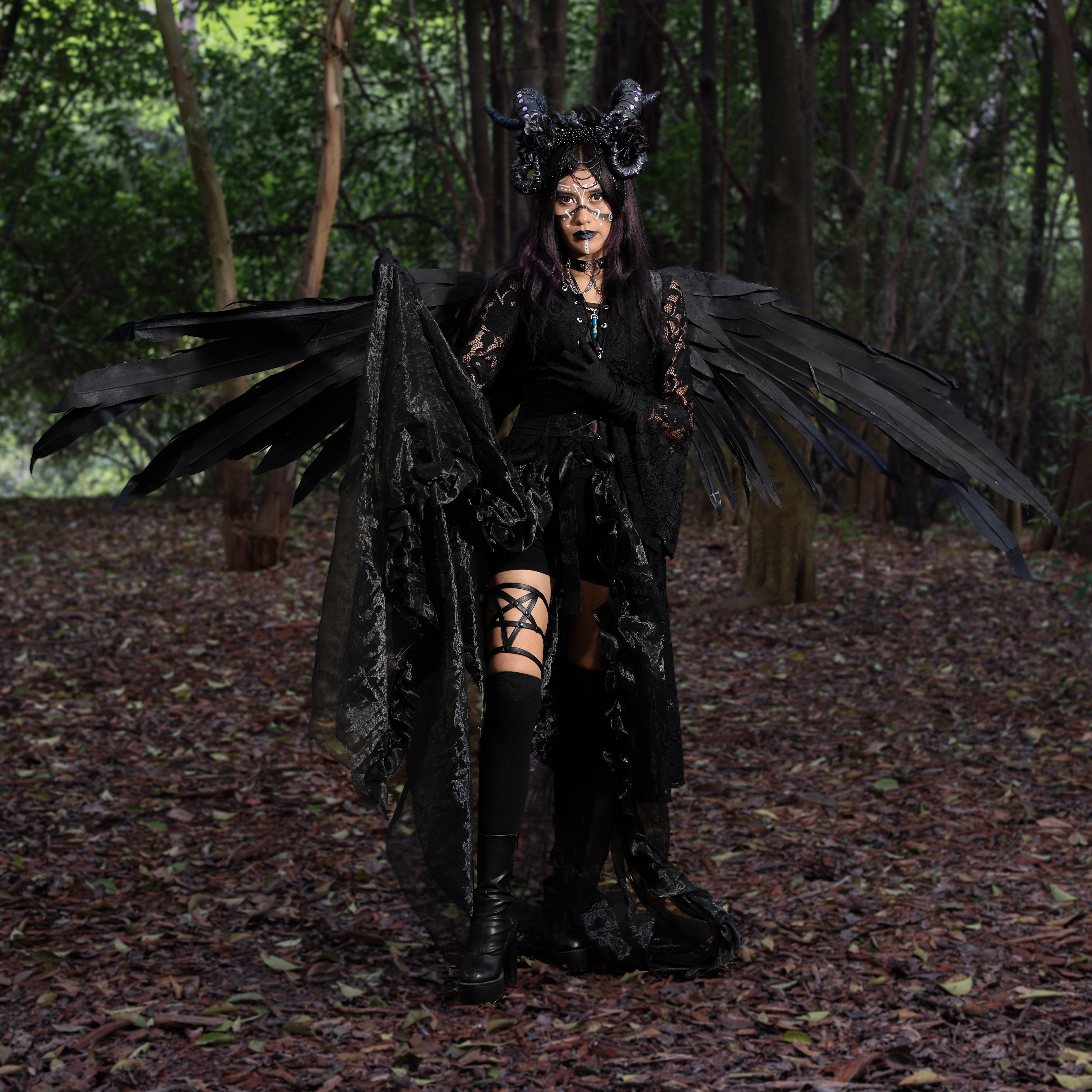 Woman in a dark, mystical costume with wings and horns posing in a forest setting.