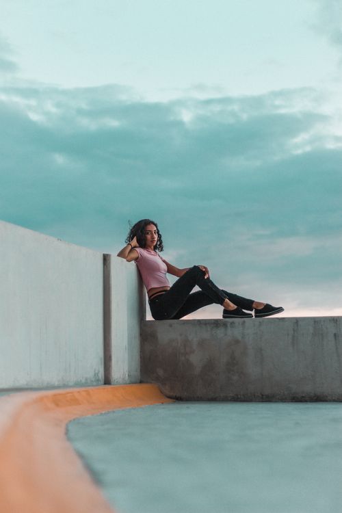Free A woman in casual attire sits relaxed on a rooftop ledge under a clear sky. Stock Photo