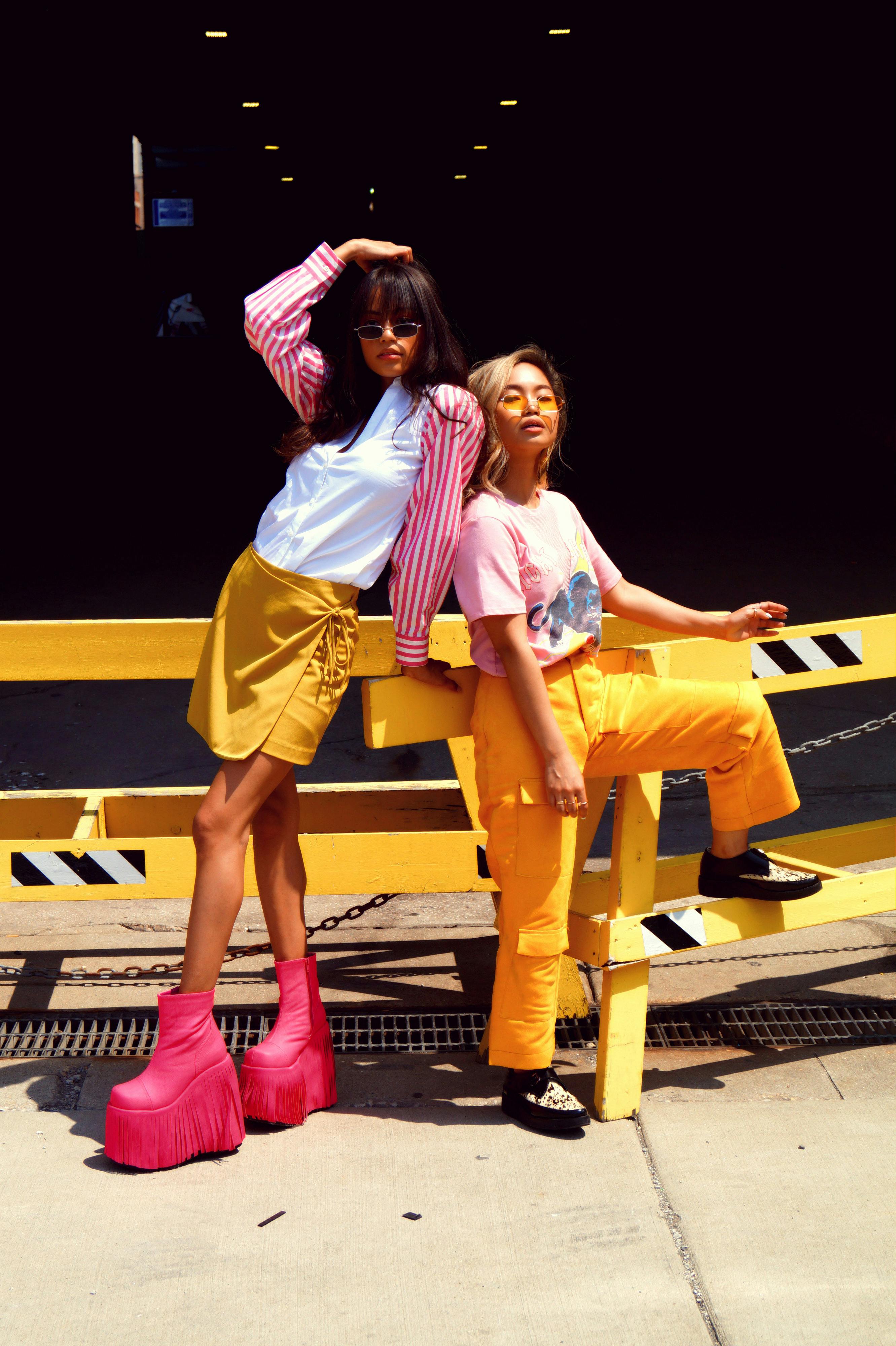 woman in pink t shirt and yellow pants next to woman in white shirt with pink and white stripe blazer