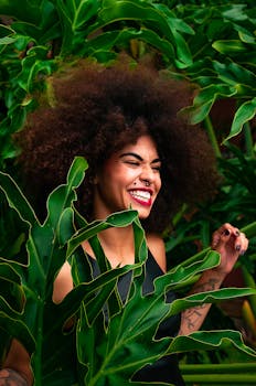一位卷发女子在郁郁葱葱的绿植间快乐地灿烂微笑，尽显自然之美与幸福之感