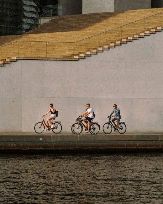 Three cyclists enjoy a summer ride along a canal in urban Berlin, framed by modern architecture.