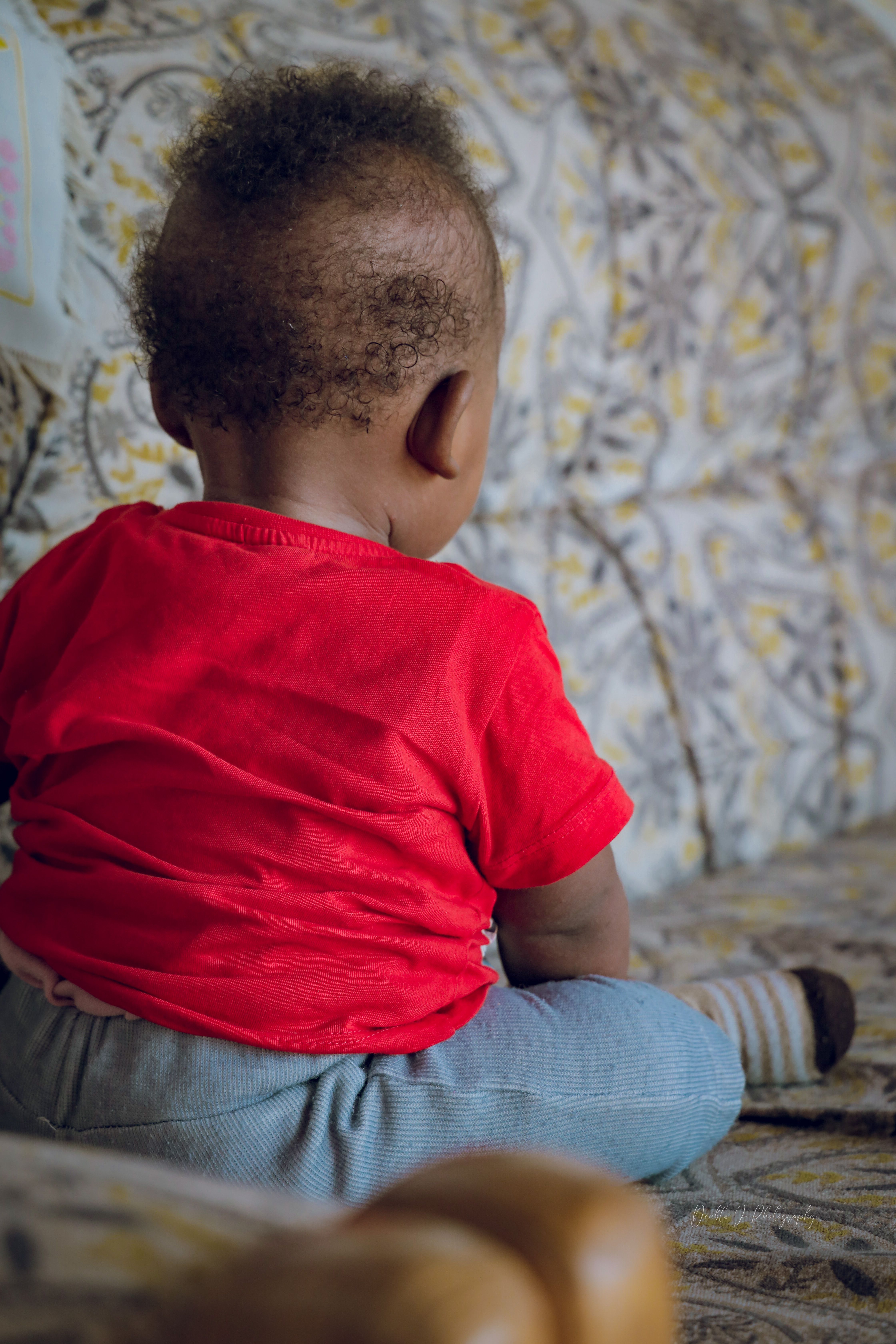 Back view of a toddler in a red t-shirt sitting on a floral-patterned couch indoors.