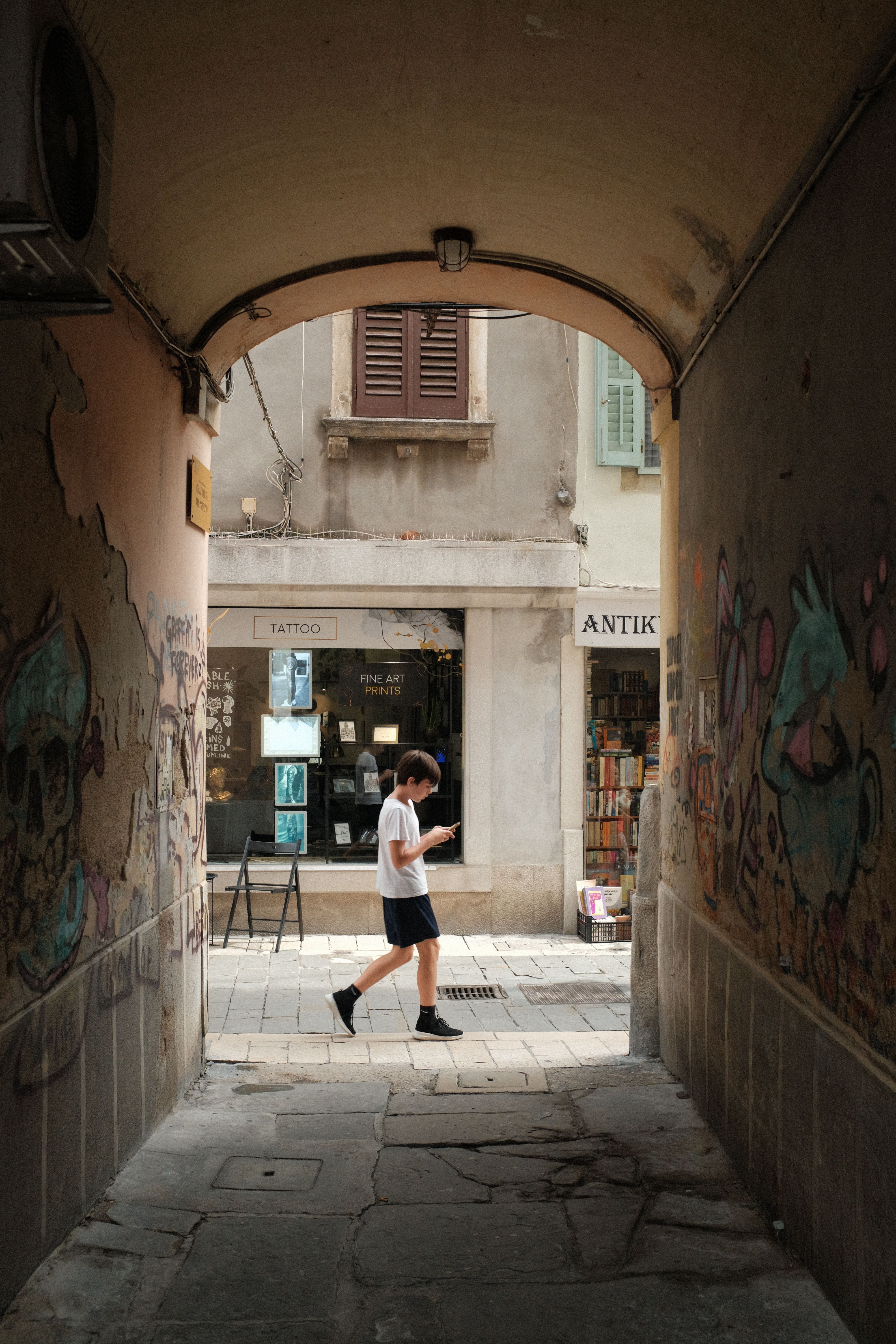 A pedestrian walks through a graffiti-lined urban passageway on a summer day.