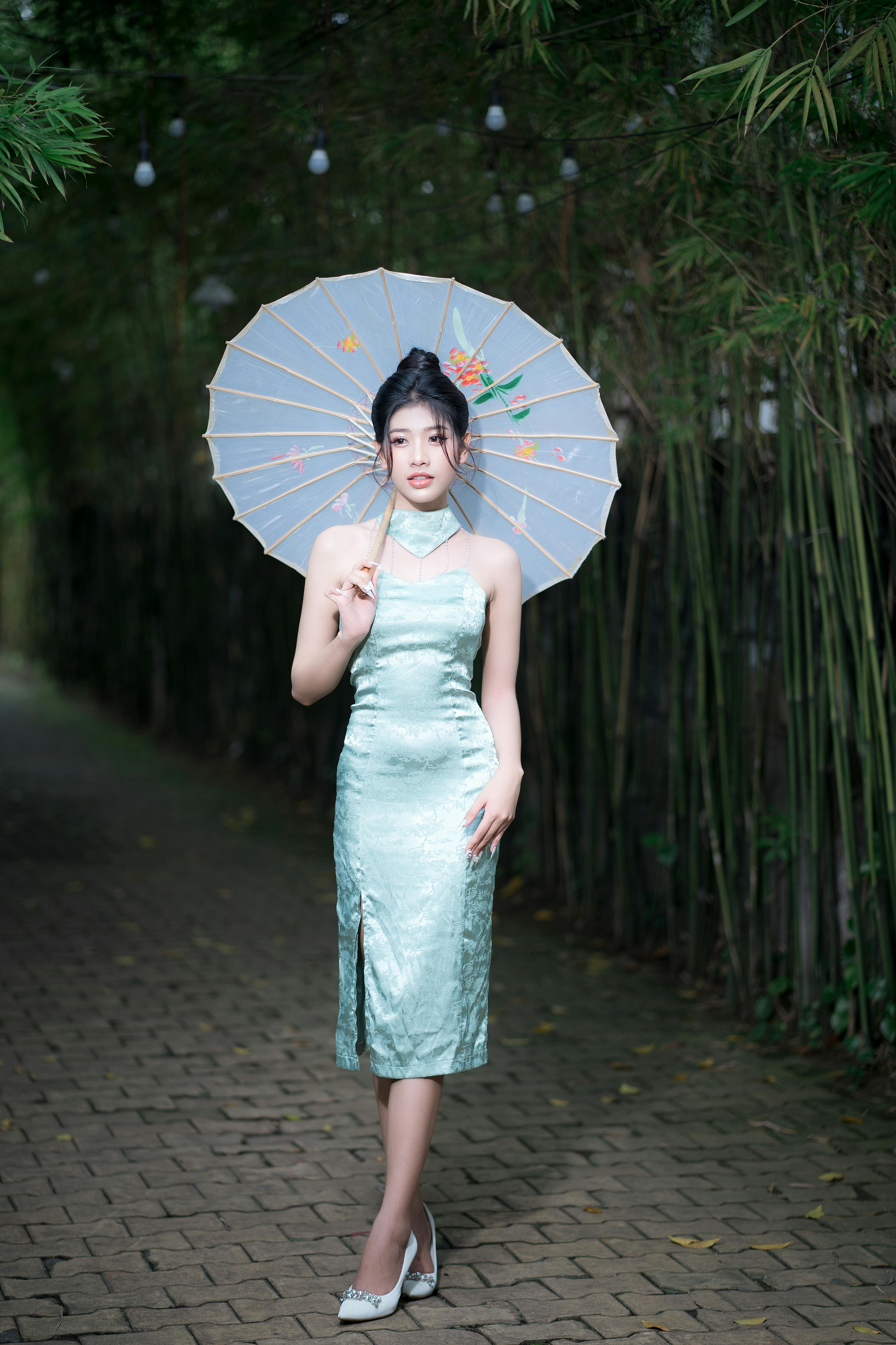 Asian woman in elegant dress posing with an umbrella in a serene outdoor setting.