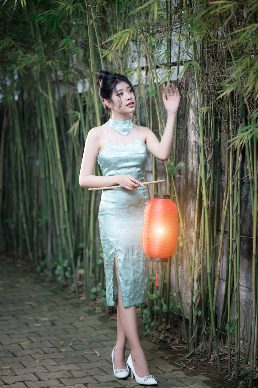 A woman in a stylish dress stands holding a paper lantern in a serene bamboo grove at dusk.