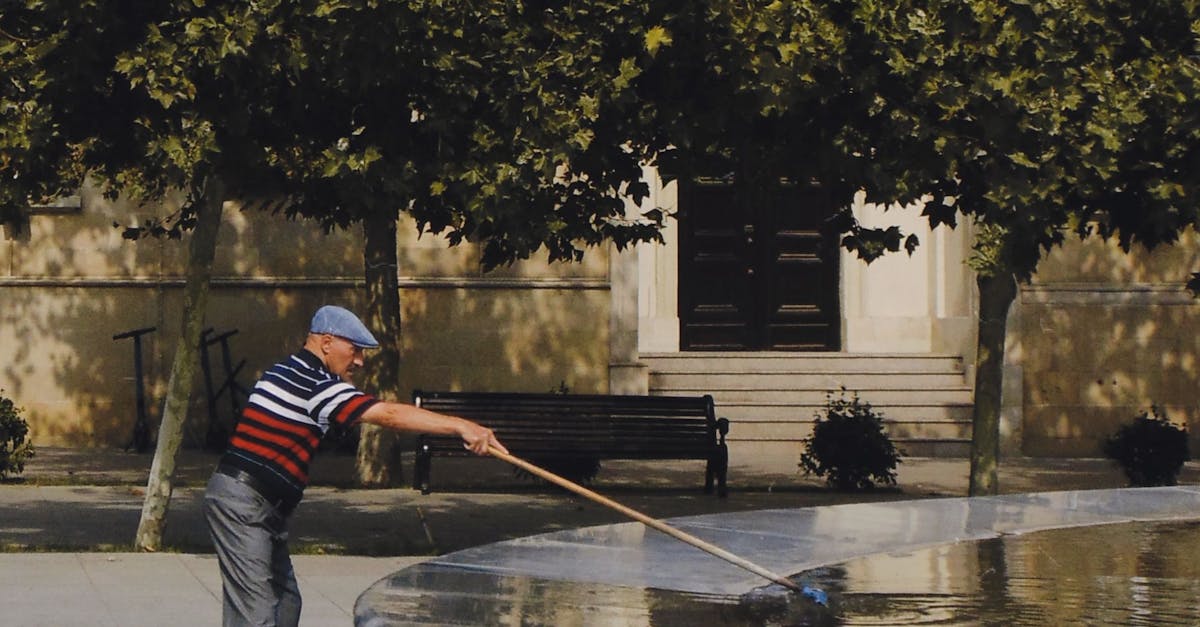 A senior man in casual clothing cleaning a fountain with a long brush outdoors.