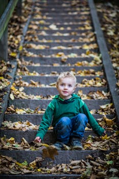 一个穿着绿色连帽衫的小男孩，坐在户外铺满秋叶的楼梯上