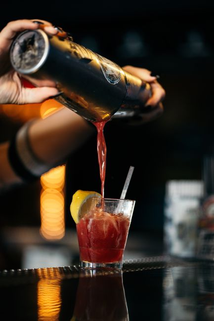 A bartender pours a vibrant red cocktail with a lemon garnish into a glass at an upscale bar.