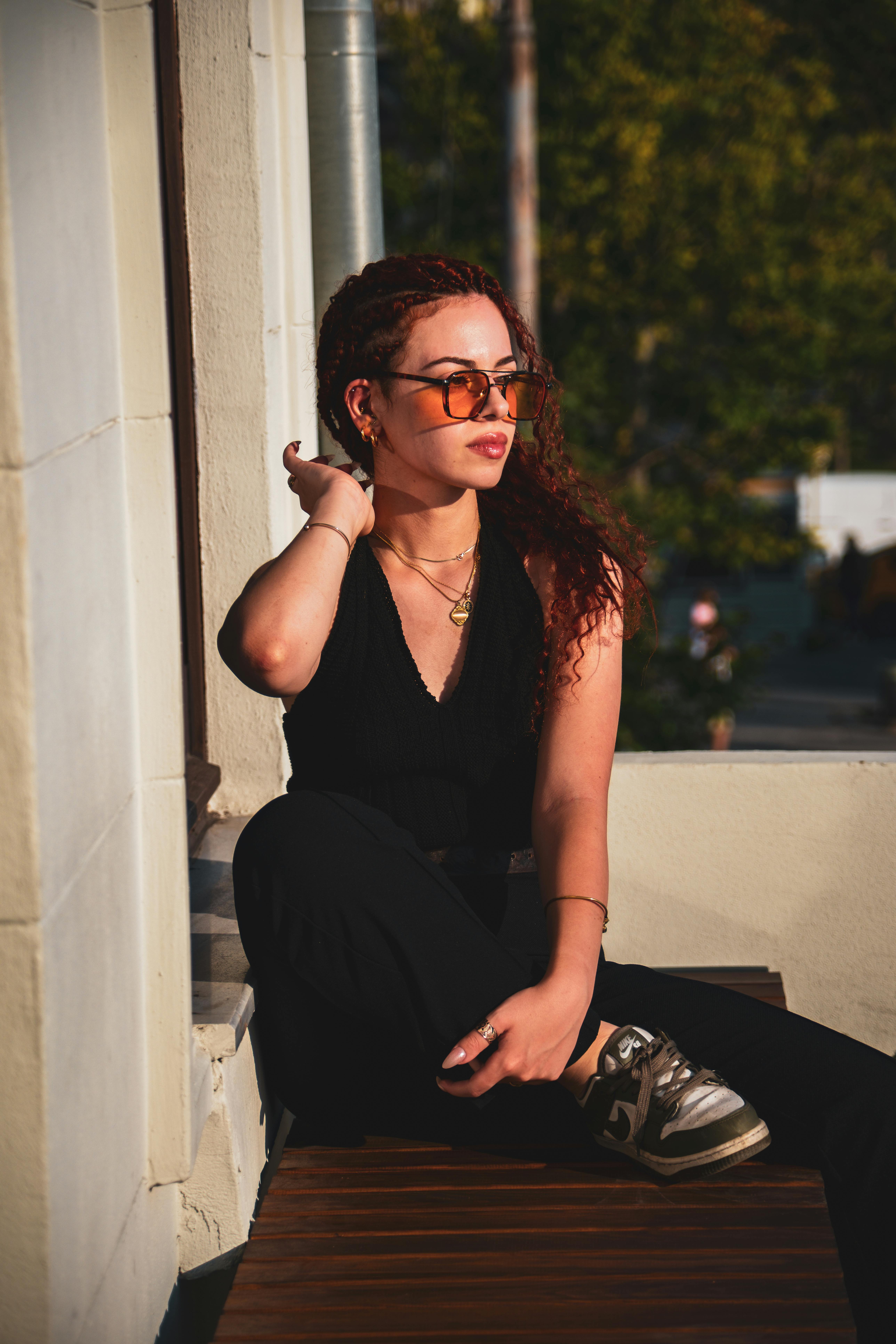 Free Elegant redhead woman in sunglasses sitting on a bench in the sun, exuding modern fashion vibes. Stock Photo