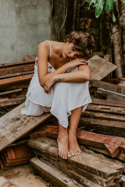 Young woman in a white dress sits thoughtfully on wood, amidst rustic surroundings.