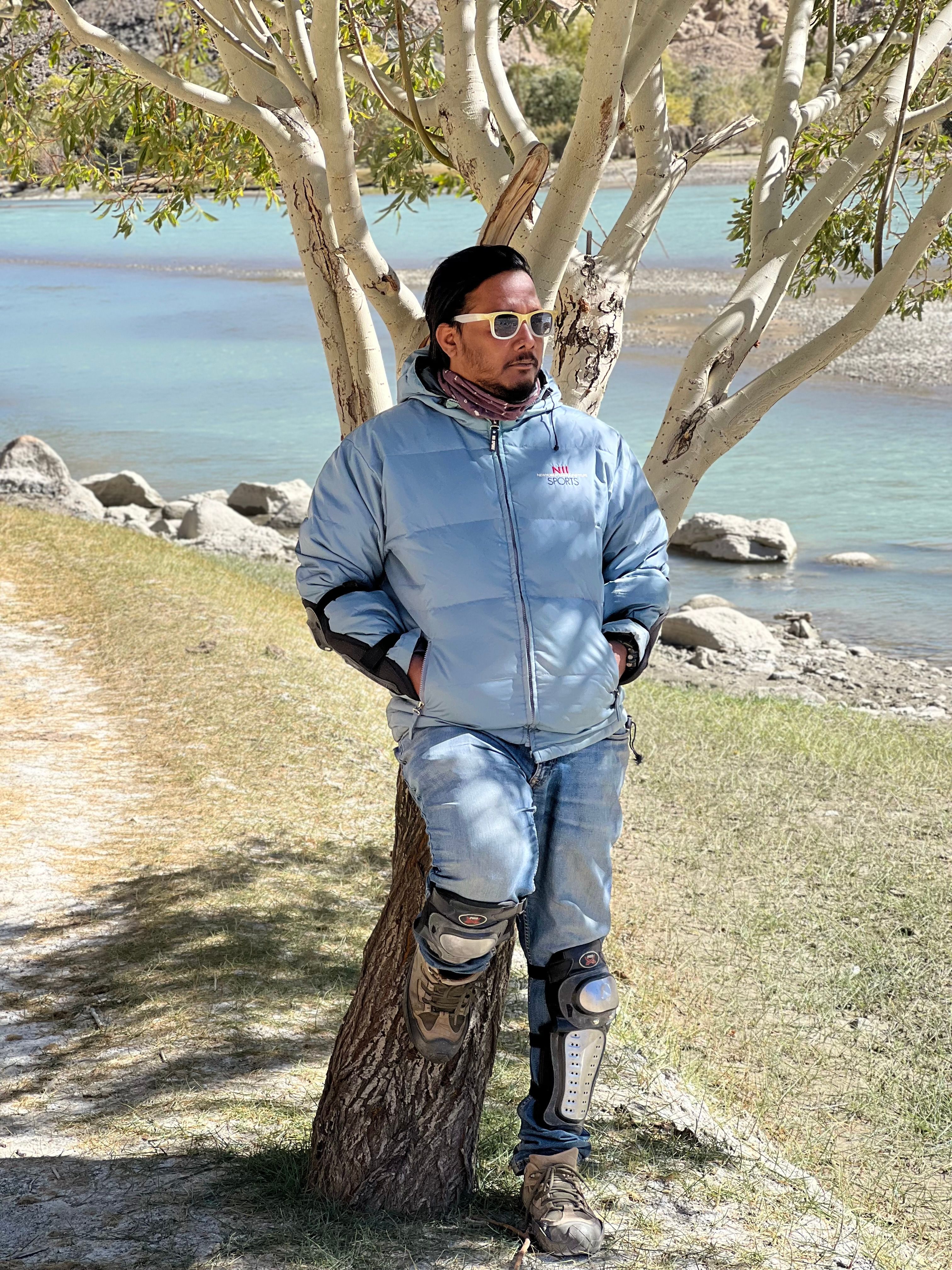 Casually dressed man in winter gear with sunglasses leaning against a tree by a serene river.