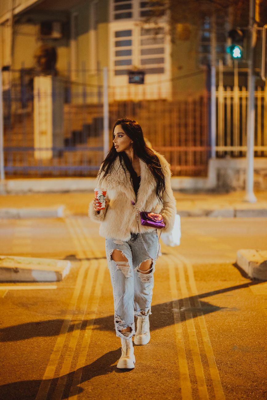 Stylish woman in a fur coat and ripped jeans walking city streets at night.