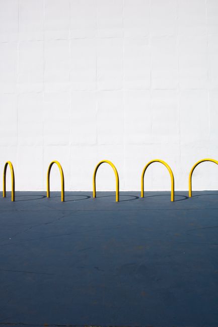 A minimalist image featuring yellow bike racks casting shadows on a blue pavement against a white wall.