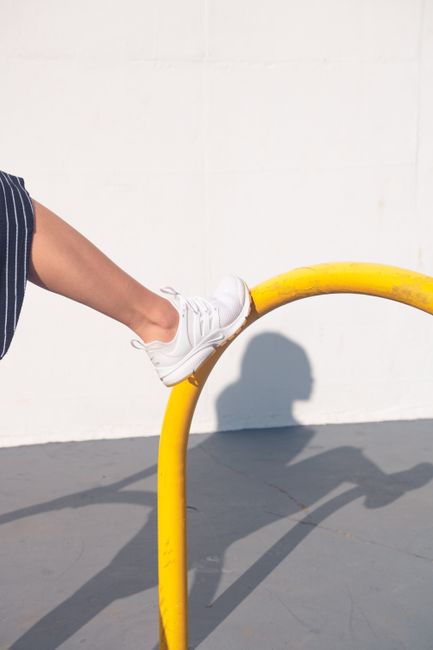 A close-up of a foot wearing a white sneaker balancing on a yellow rail in bright light, casting a shadow.