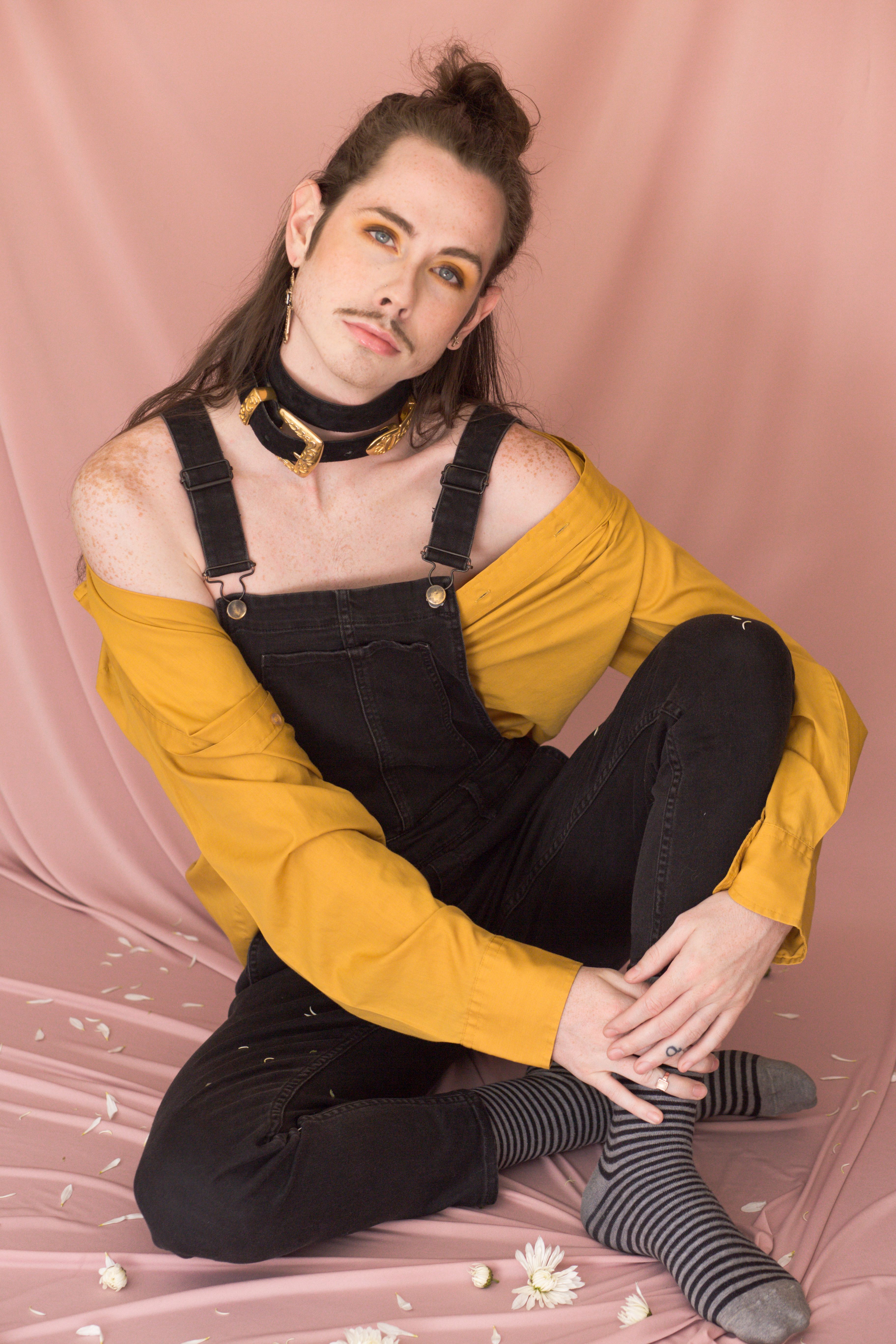 Elegant portrait of a young adult in black overalls and mustard shirt, sitting against a pink backdrop.