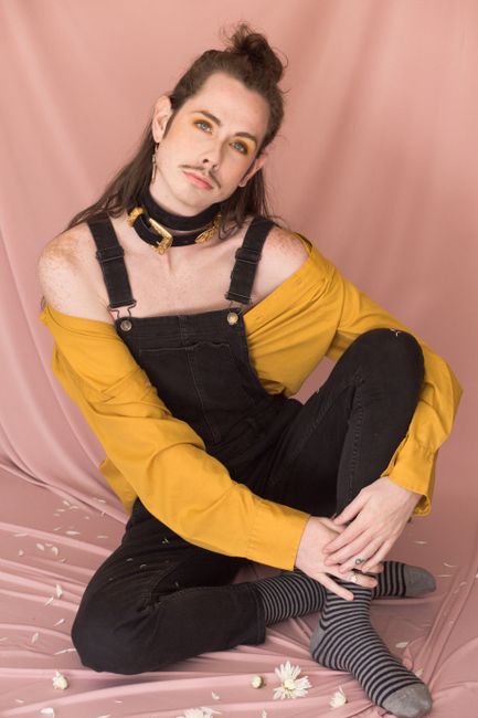 Elegant portrait of a young adult in black overalls and mustard shirt, sitting against a pink backdrop.