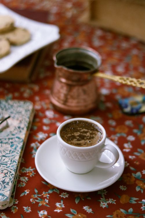 Free A warm Turkish coffee served in a white cup on floral tablecloth with a copper cezve in the background. Stock Photo