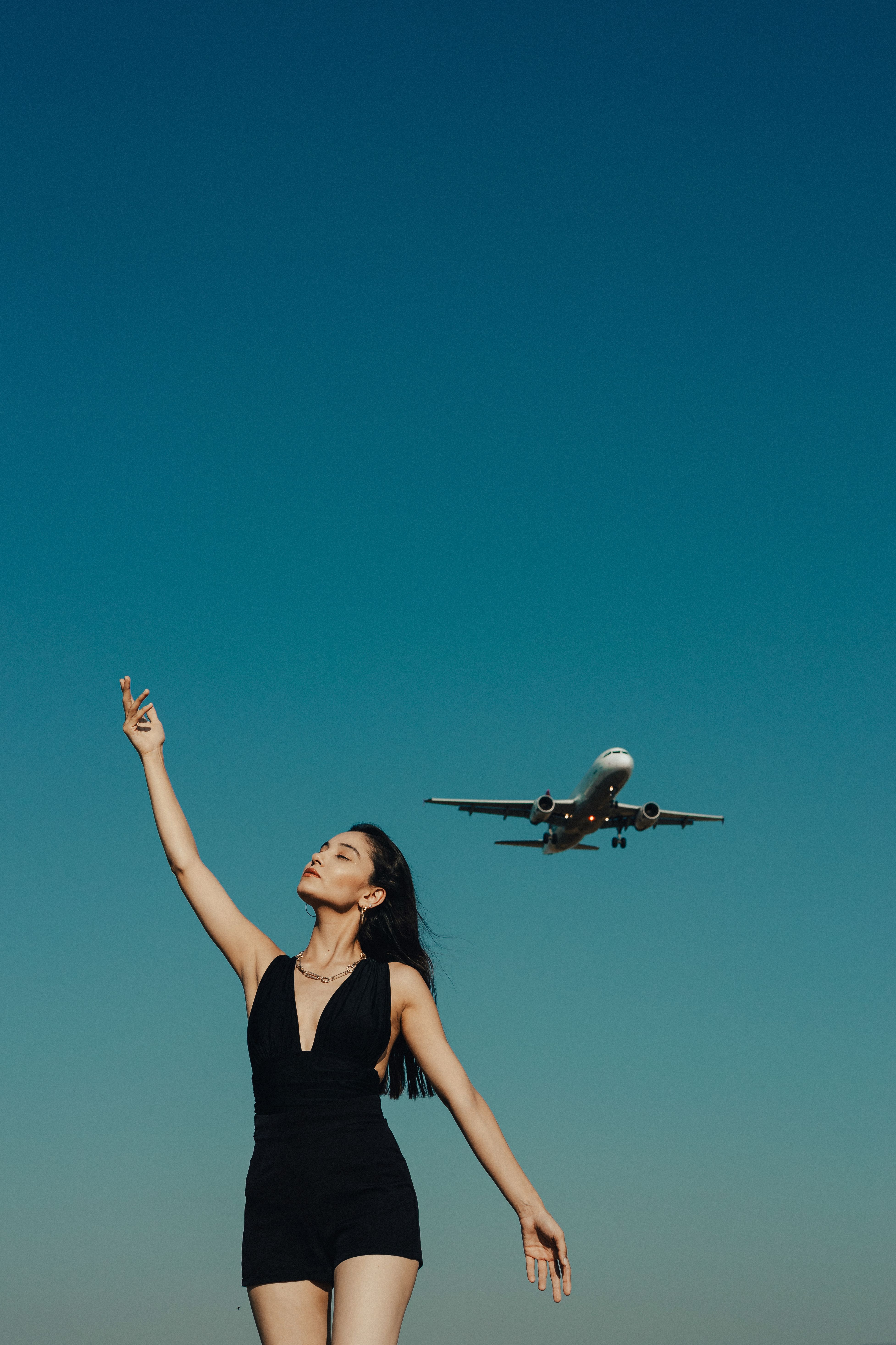 A carefree young woman raises her arm joyfully as an airplane flies overhead against a clear blue summer sky.
