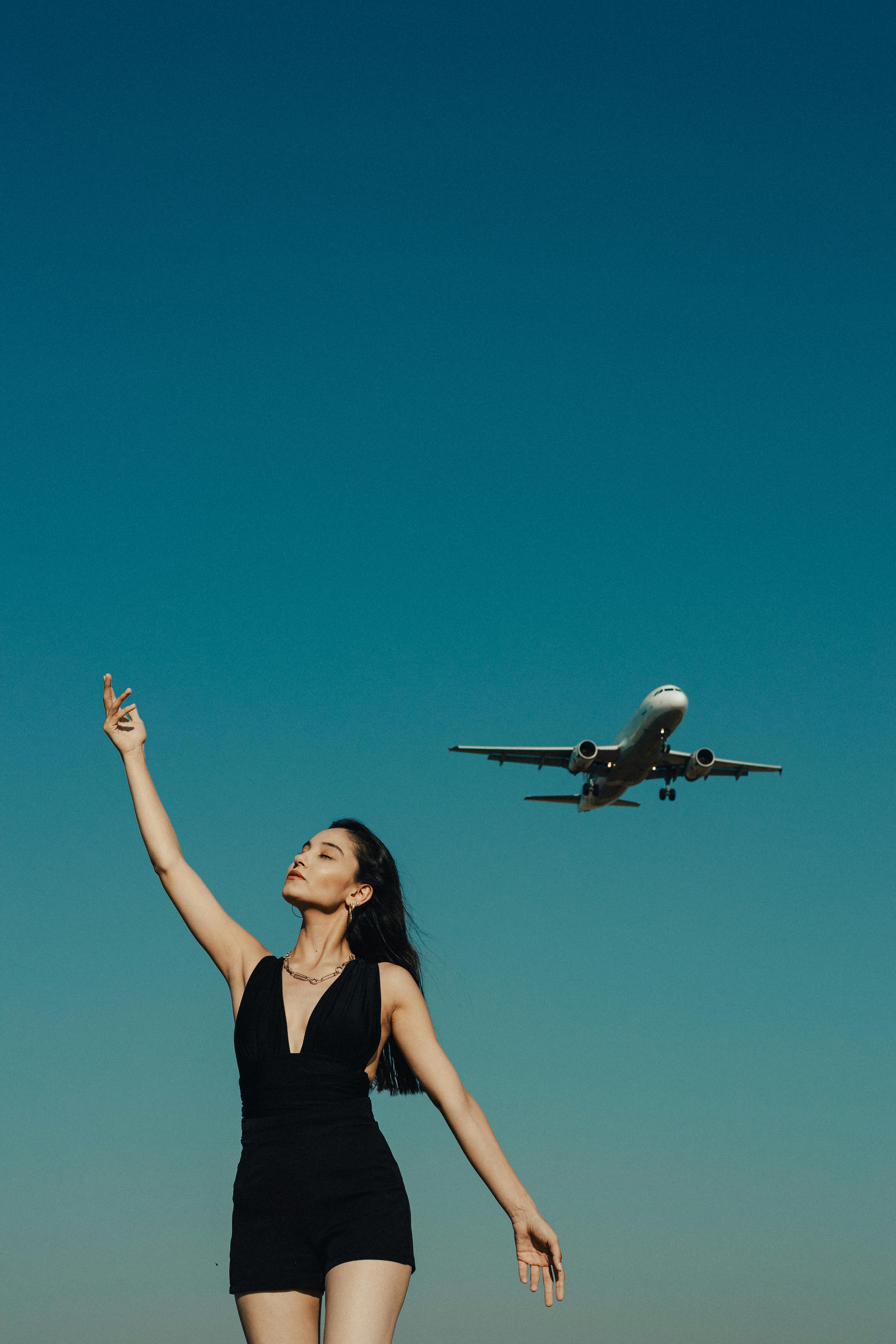 Woman raising hand under a blue sky as an airplane flies above, evoking freedom and travel dreams.