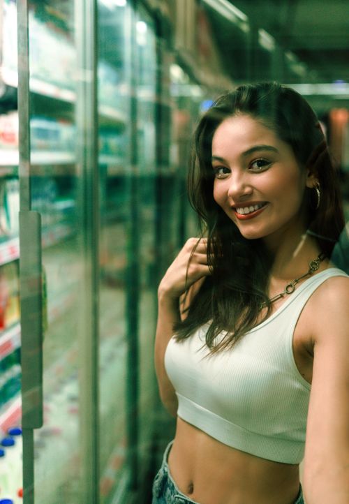 Free Young woman enjoying a relaxed shopping experience in a grocery store aisle, wearing casual summer attire. Stock Photo