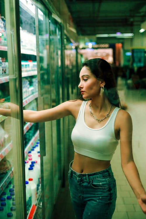 A woman in a white top and jeans is looking at a refrigerator