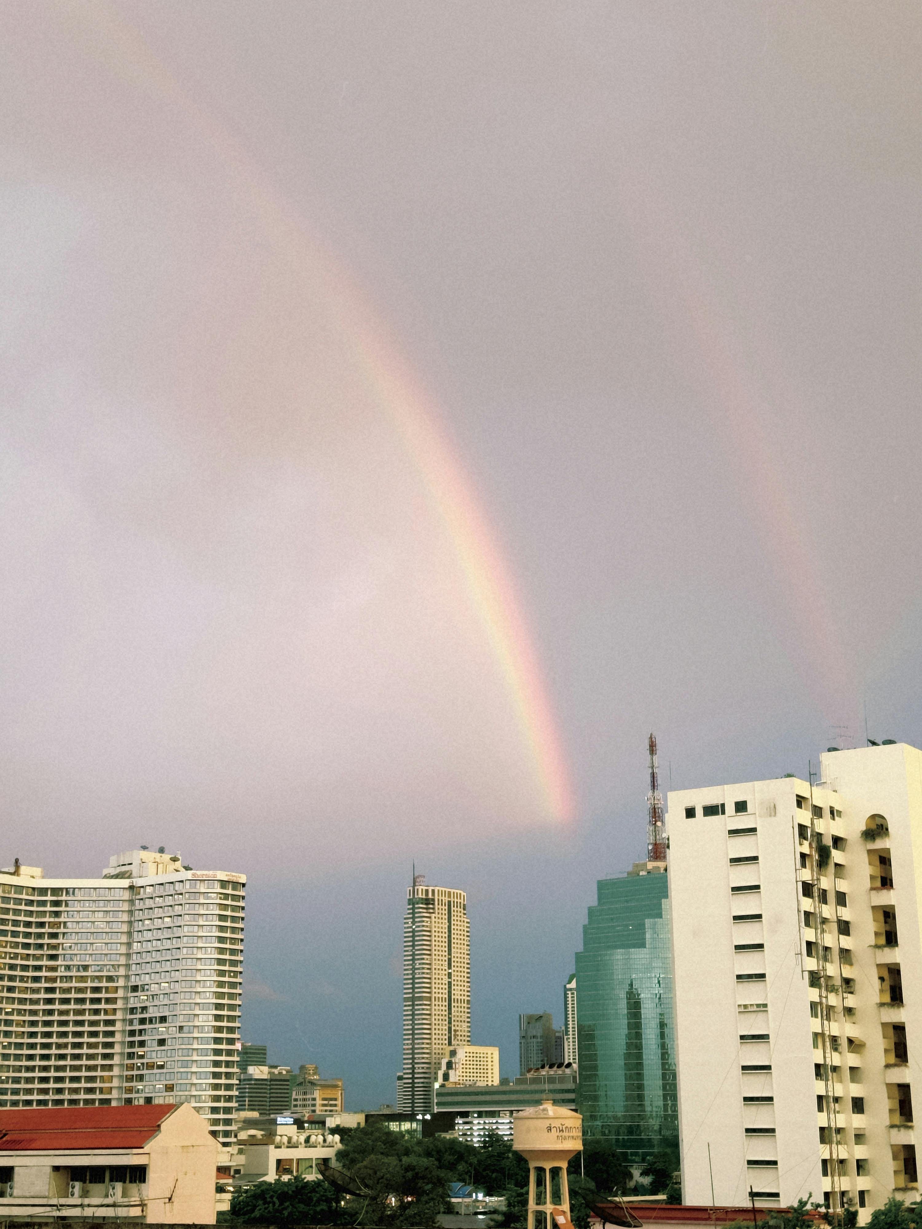 A colorful rainbow arches over urban skyline amidst cloudy sky.