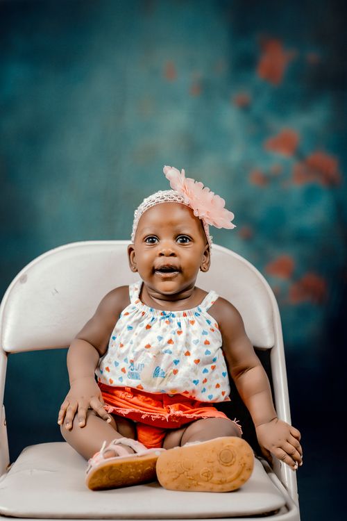 Free Charming portrait of a baby on a chair with a pink floral headband and colorful outfit. Stock Photo