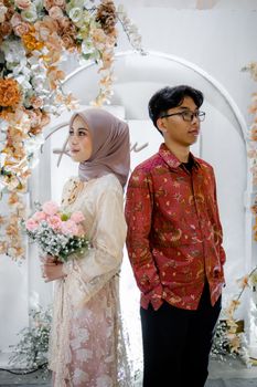 A couple in traditional attire poses elegantly under a floral arch, embodying love and tradition. - Fotografer Sidoarjo