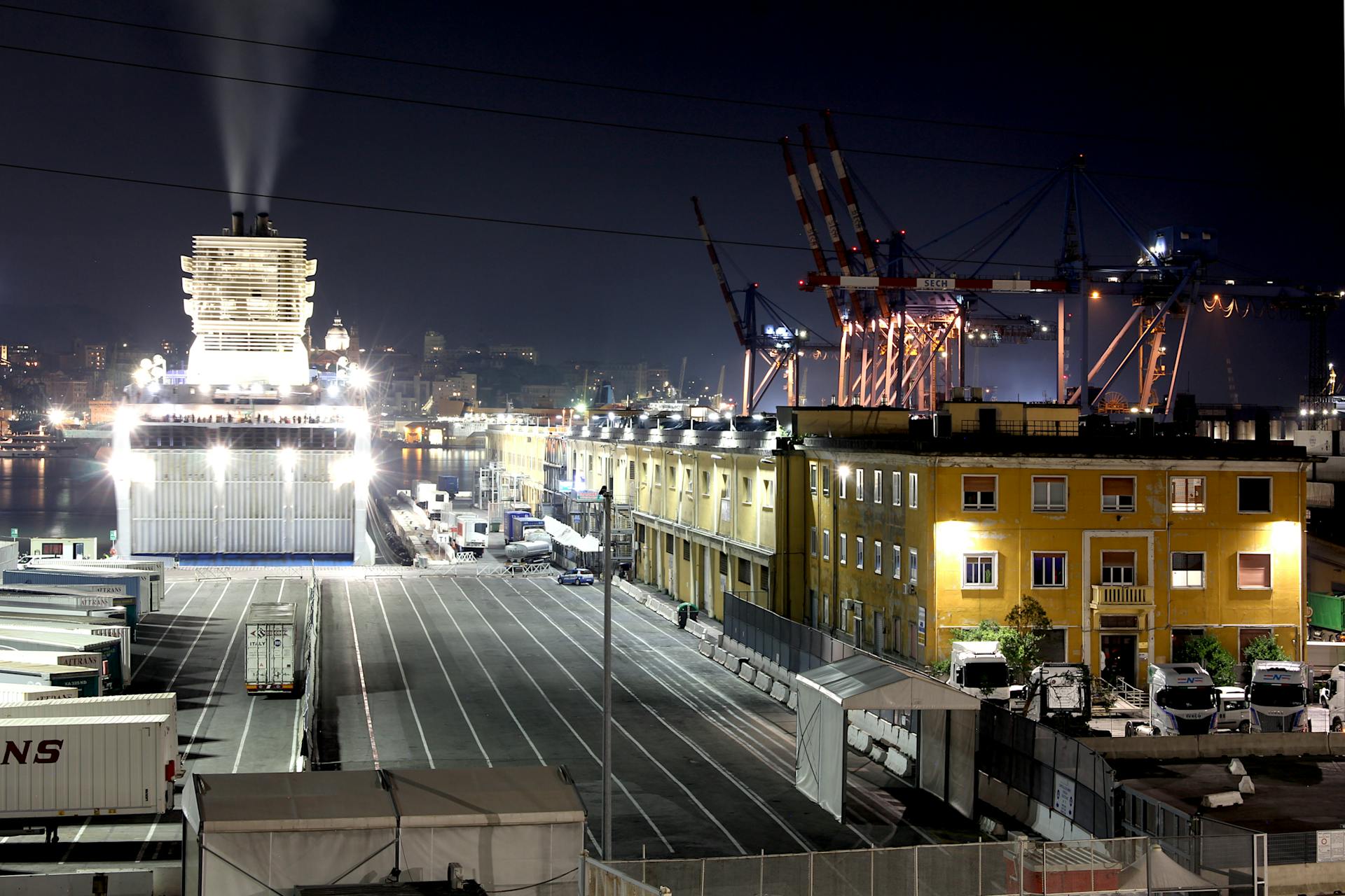 A bustling night scene at Genoa harbor showcasing a docked ship, industrial cranes, and lit buildings.