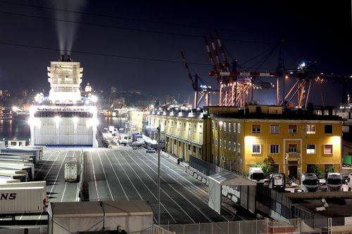 Free A bustling night scene at Genoa harbor showcasing a docked ship, industrial cranes, and lit buildings. Stock Photo