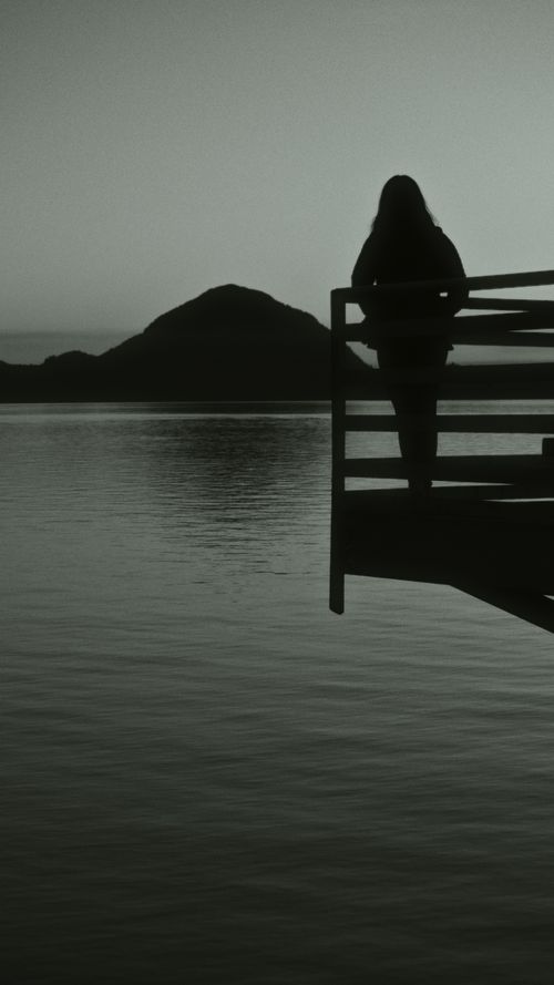 Free A serene silhouette of a person standing on a pier by the lake, with mountains in the background at dusk. Stock Photo