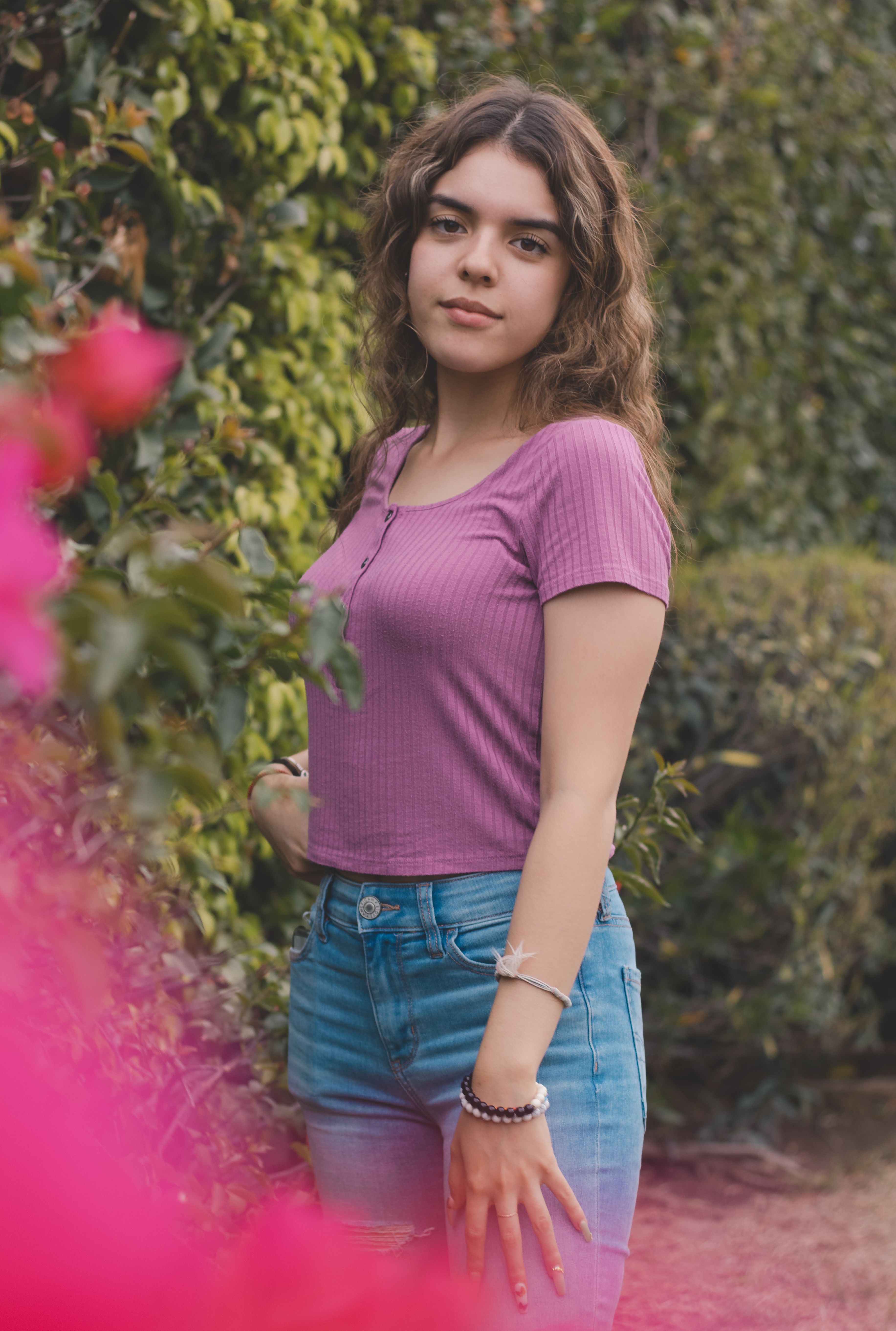 Portrait of a young woman in a garden, wearing a purple top and jeans, surrounded by greenery.