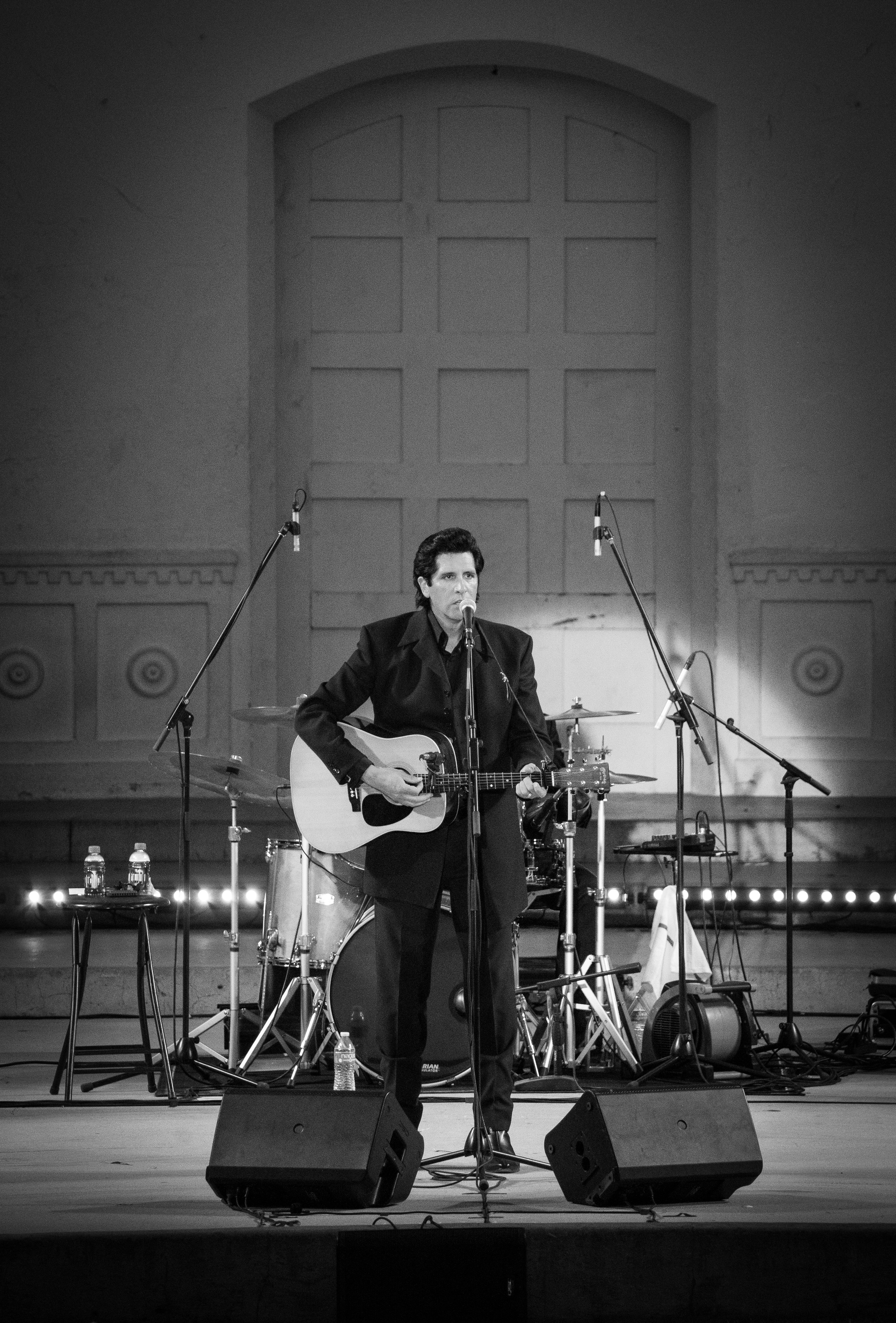 Black and white photo of a musician performing with guitar and microphones on stage.