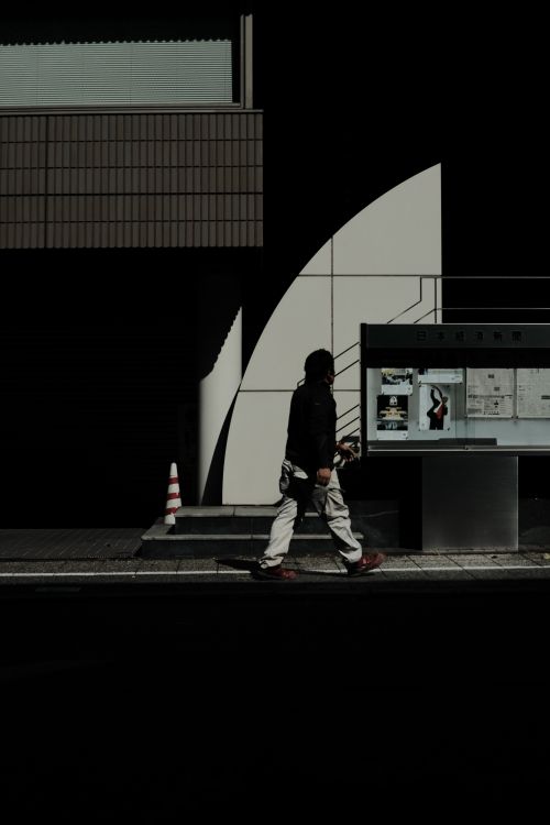 Free A solitary pedestrian walks along an urban sidewalk, casting a shadow on modern architecture. Stock Photo