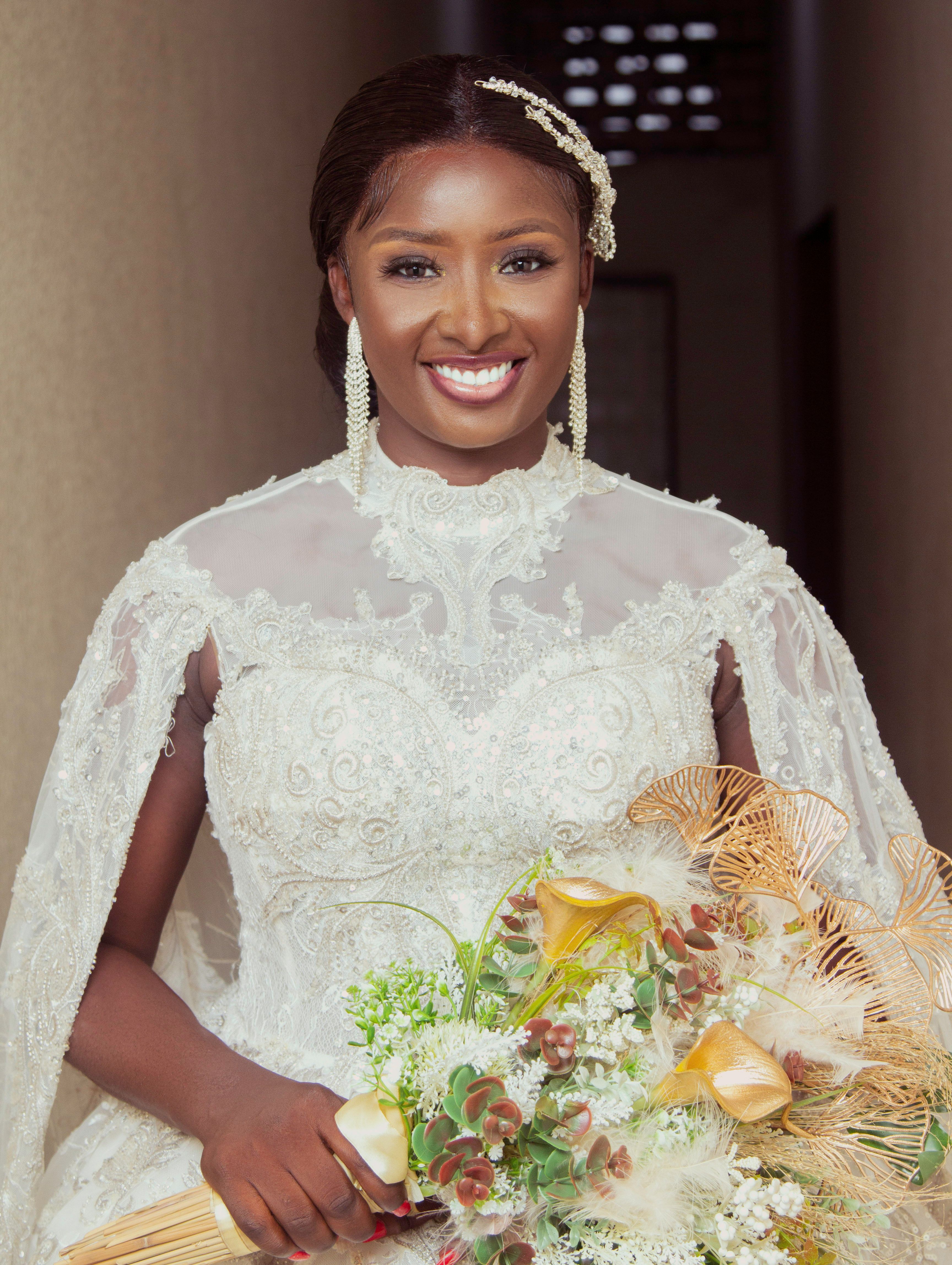 Elegant bride in lace gown smiling indoors, holding colorful bouquet.